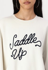 Felpa color crema con ricamo blu navy che recita "Saddle Up". Tessuto morbido con scollatura rotonda e maniche lunghe.