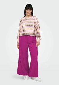 Maglione lavorato a maglia con strisce multicolori, abbinato a pantaloni a gamba larga e a costine di un brillante colore rosa. Il modella si trova davanti a uno sfondo chiaro.