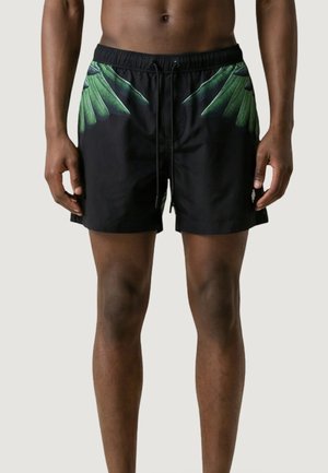 Uomo che indossa pantaloncini da bagno neri con disegni di foglie di palma verdi vicino alla vita, in piedi contro uno sfondo semplice.