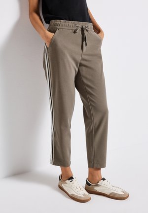 Personne portant un pantalon marron fuselé avec des rayures latérales blanches et noires et des baskets blanches, se tenant contre un mur blanc.