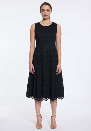 Vrouw staand, gekleed in een mouwloze zwarte kanten midi-jurk met bloemenpatronen en gouden sandalen met hoge hakken en enkelriem op een witte achtergrond.