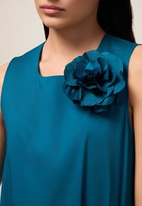 Teal mouwloos jurkje met een vierkante halslijn, voorzien van een grote getextureerde bloemaccent op de linkerschouder. Soepele stof.