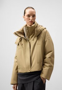 Beżowa wodoodporna kurtka z wysokim kołnierzem, dużymi kieszeniami i sznurkiem; ma teksturowane akcenty przy dolnej krawędzi oraz logo.