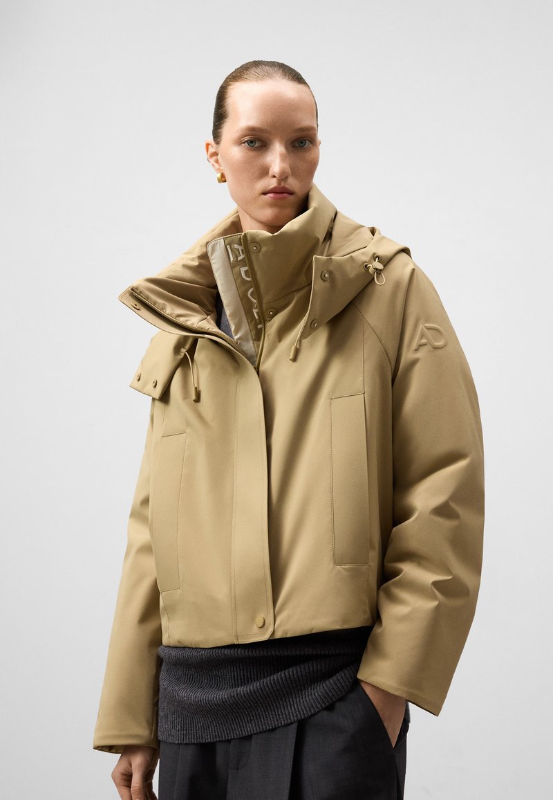 Beżowa wodoodporna kurtka z wysokim kołnierzem, dużymi kieszeniami i sznurkiem; ma teksturowane akcenty przy dolnej krawędzi oraz logo.
