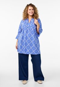 Blauwe tuniek met een geometrisch wit patroon, lange mouwen en een gevorkte kraag, gecombineerd met marineblauwe wijde broek en lichte schoenen.