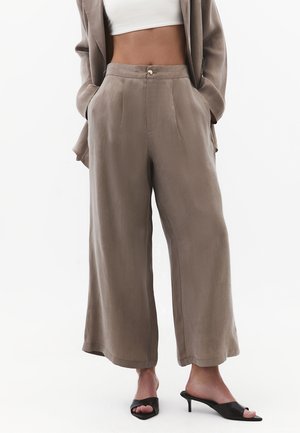 Donna che indossa pantaloni beige a vita alta, larghi, con tasche frontali, abbinati a sabot neri con tacco e punta aperta, top bianco corto e blazer coordinato.