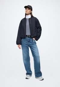 Tamsiai mėlyna bombonešio striukė dėvima ant pilko megztinio, derinama su šviesiai mėlynais plačiomis kojomis džinsais ir pilkais sportiniais bateliais. Aksesuarai: tamsi beisbolo kepuraitė.