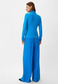 Niebieska bluzka z długim rękawem i szerokie spodnie, obie z gładkiego materiału. Bluzka ma stójkę, a spodnie są luźne i mają smukły kształt.
