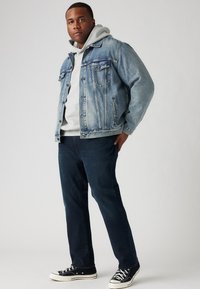 Lichtblauwe spijkerjack over een grijze hoodie, gecombineerd met donkerblauwe jeans en zwarte sneakers. Het jack heeft voorzakken en een knoopsluiting.