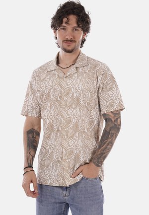 Uomo con capelli ricci che indossa una camicia beige a maniche corte con motivi e jeans, mostrando tatuaggi su entrambi gli avambracci, posando con una mano in tasca.