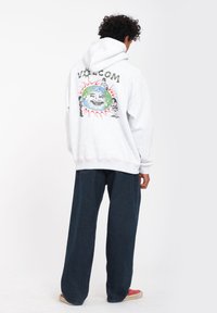 Grijze hoodie met grote grafische print op de achterkant met een globe-design en figuren, gecombineerd met donkere wijde broekspijpen en rode schoenen.