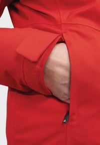 Rote Jacke mit strukturiertem Kragen und mit Klettverschluss verstellbaren Bündchen. In der Seiten Tasche ist eine Hand sichtbar, mit einem Reißverschlussdetail.
