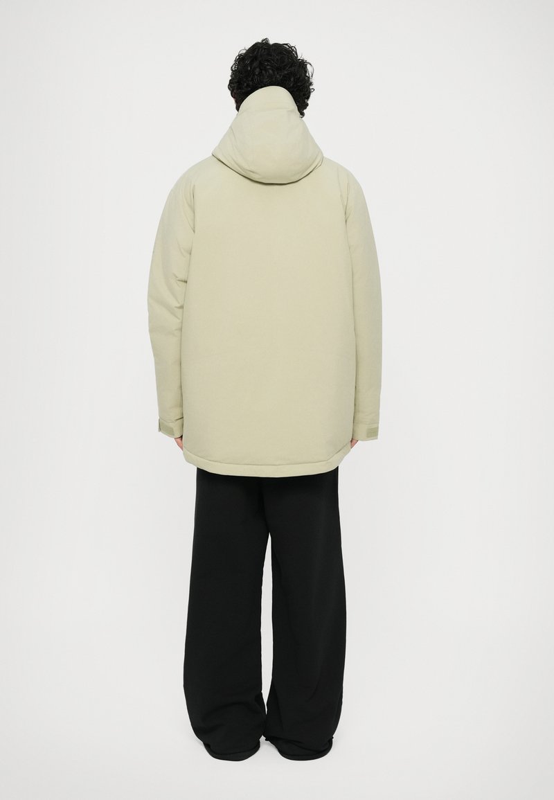 Veste à capuche de couleur vert clair avec une texture douce, vue de l'arrière, associée à un pantalon noir ample. Design simple, sans matériel visible.