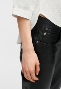 Κοντινή λήψη του χεριού ενός ατόμου που ακουμπά κοντά στην εμπρόσθια τσέπη μαύρου δερμάτινου παντελονιού, φορώντας λευκό υφασμάτινο μπλουζάκι με μακριά μανίκια.