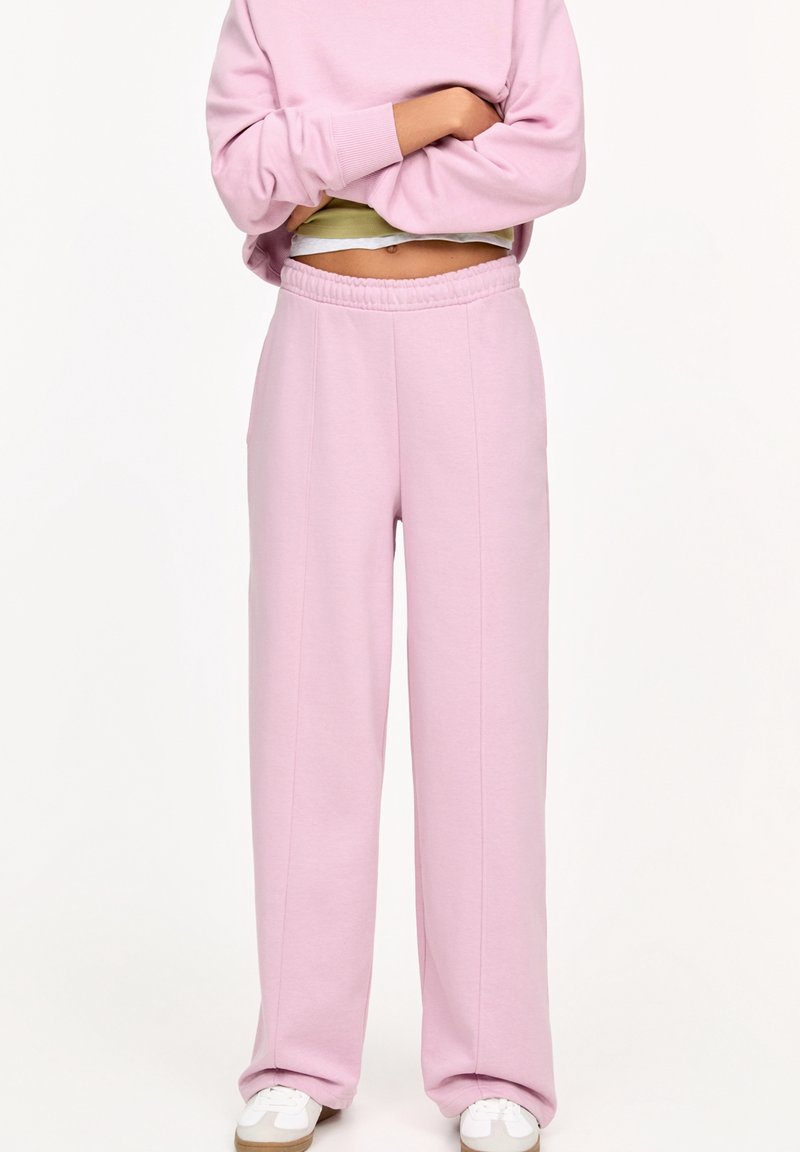 Růžové tepláky vyrobené z měkké tkaniny, s volným střihem, elastickým pasem a bočními kapsami. Doplněné o bílé tenisky.