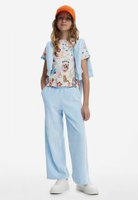 Hellblaue Hosen mit weitem Bein, kombiniert mit einem kurzärmeligen T-Shirt mit bunten Tiermotiven. Orangefarbene Mütze und leichter Pullover lässig darüber gelegt.