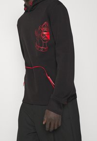 Schwarzer Hoodie mit einer roten Linienzeichnung eines Hundehaars, Reißverschlussakzenten und verstellbaren roten Kordeln an der Kapuze. Weicher Stoff.