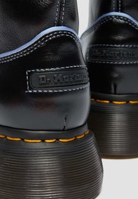 Μαύρα δερμάτινα μποτάκια Dr. Martens με κίτρινο ράψιμο, χοντρές ραβδωτές σόλες και ανάγλυφες λογότυπες ετικέτες στα πίσω τακούνια.