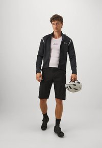 Svart sykkeljakke med reflekterende detaljer, hvit kortermet topp, svarte shorts og sykkelhjelm i hånden. Hurtigtørkende stoff egnet for sport.