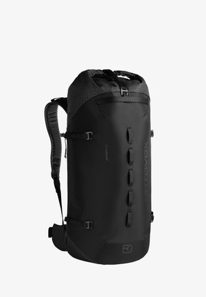 Mann trägt ein graues T-Shirt, hellbraune Hose, Wanderschuhe und einen großen schwarzen Wanderrucksack, steht vor einem einfarbigen Hintergrund.