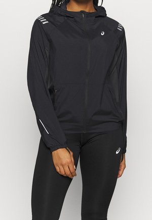 Personne portant une veste à capuche noire zippée et des leggings noirs avec un logo blanc sur la poitrine, la manche et la cuisse, debout devant un fond uni.