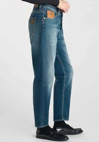 Jeans de mezclilla azul con corte recto, desgaste sutil, parche de cuero con logotipo y herrajes de latón, combinados con mocasines negros y calcetines.