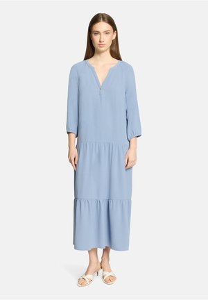 Frau steht und trägt ein hellblaues, langärmeliges, gestuftes Midikleid und weiße Sandalen mit Absatz vor einem schlichten weißen Hintergrund.