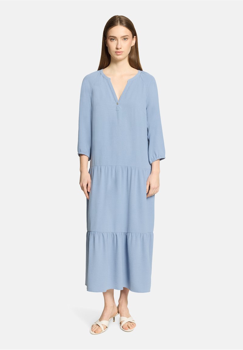 Femme debout, portant une robe midi à manches longues bleu clair à volants et des sandales blanches à talons, sur un fond blanc uni.