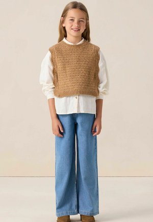 Bruine gebreide cropped vest over een witte blouse, gecombineerd met wijde blauwe jeans en bruine enkellaarzen. Soepele texturen en gelaagd ontwerp.