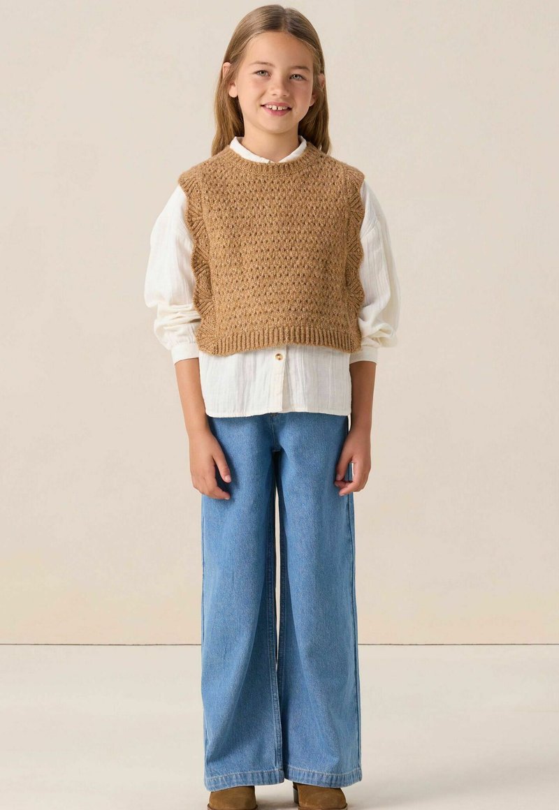 Un gilet court en tricot marron porté sur une chemise blanche à boutons, associé à un jean bleu à jambes larges et des bottines marron. Textures lisses et design superposé.