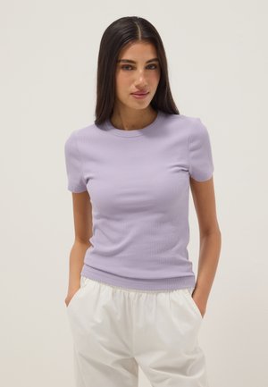 Mujer joven con cabello largo y oscuro, vestida con una camiseta ajustada de manga corta color lavanda y pantalones blancos con las manos en los bolsillos.