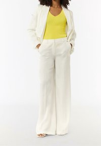 Cremfarbener Blazer über einem gelben gerippten Tanktop, kombiniert mit weißen weitem Hosen. Glatte Textur, maßgeschneiderte Passform und klare Linien.
