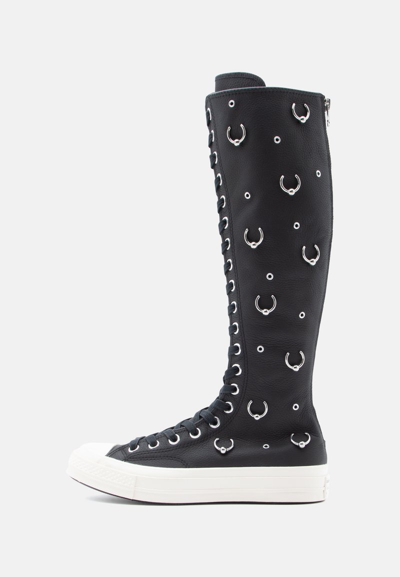 Botas de caneira alta pretas com atacadores em pele texturizada, apresentando detalhes em metal prateado e ilhós circulares, com uma sola em borracha branca.