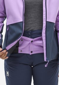 Veste de ski multicolore en violet et bleu marine avec une fermeture éclair ; associée à un pantalon de neige bleu marine. Présente une fermeture à pression et des détails de logo.