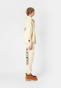 Joven con sudadera beige y pantalones con bolsillo lateral y cinta colgante, vistiendo botas marrones de cordones, de pie frente a un fondo blanco.