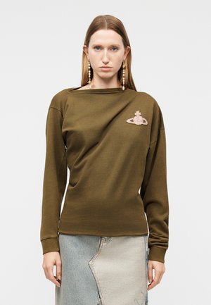 Egyenes hajú nő olívazöld hosszú ujjú felsőt visel logós címkével és kéttónusú farmer szoknyát, egyszerű háttér előtt áll.