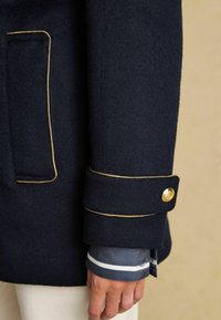 Tumesinine villane jakk, mille varrukal on kuldne trimm ja nööpide detail, all on kihiline helesinine särk valgete triipudega.