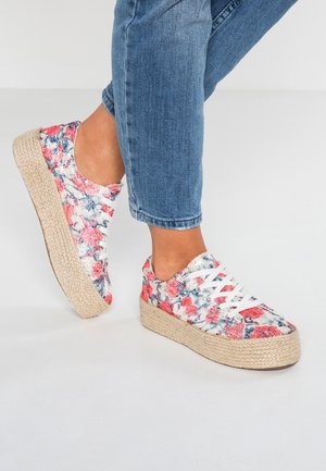 Fødder iført blå jeans og platåsneakers med blomsterprint, hvide snørebånd og flettede rejsåler mod en ensfarvet baggrund.