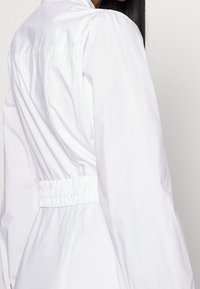 Camicia bianca a maniche lunghe con vita aderente, tessuto texturizzato e spalle a palloncino. Presenta un bordo elastico arricciato in vita.