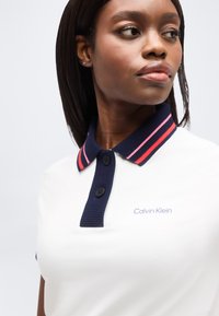 Γυναίκα με σκούρο δέρμα και ίσια μαλλιά που φοράει λευκό πόλο πουκάμισο με σκούρο μπλε γιακά και ροζ ρίγες, με το λογότυπο Calvin Klein ορατό στο στήθος.