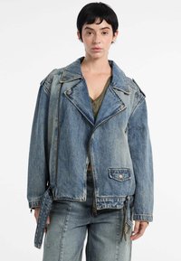 Model in een loszittende denim jas met een ritssluiting en kleppen, gestyled met een groene top en bijpassende denim jeans tegen een witte achtergrond.