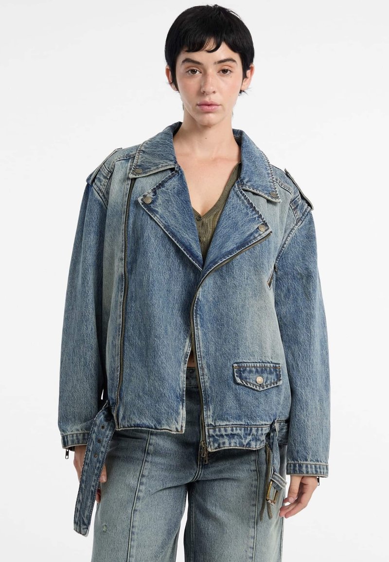Model in een loszittende denim jas met een ritssluiting en kleppen, gestyled met een groene top en bijpassende denim jeans tegen een witte achtergrond.