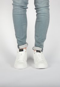 Sneakers bianchi in pelle con una texture liscia, punta rotonda e suola in gomma piatta, con logo nero sulla linguetta e jeans azzurri arrotolati.