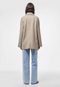 Cappotto di lana beige con colletto, vestibilità ampia e maniche larghe, abbinato a jeans di denim azzurro, visto da dietro.