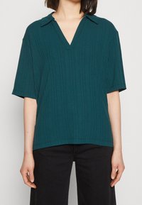 Teal ribbad kortärmad blus med V-ringning och spetsig krage. Tyget har en texturerad yta med vertikala linjer och en avslappnad passform.