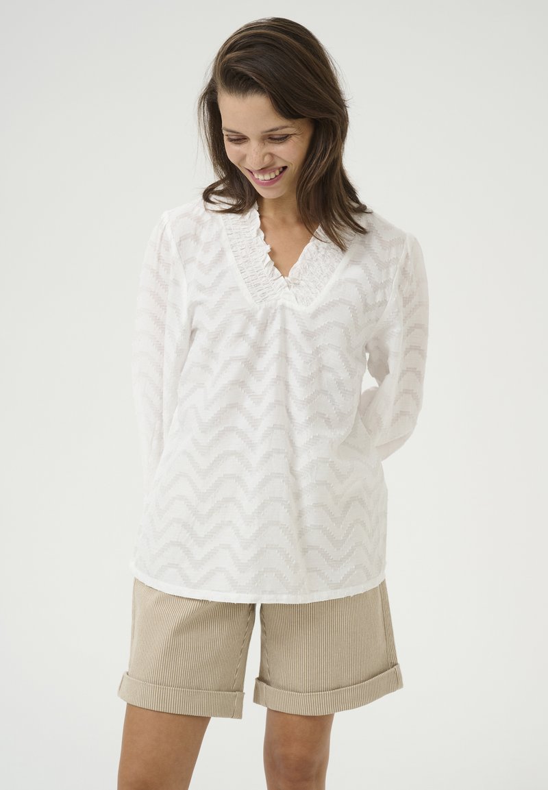Blouse blanche avec un motif de vagues texturé, décolleté en V avec détails en volants, manches longues. Associée à un short beige avec revers.