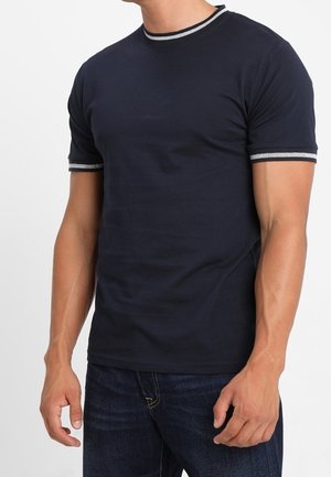 Homme portant un t-shirt bleu marine à manches courtes avec des bordures grises au niveau du col et des manches, associé à un jean bleu foncé.