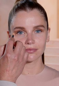 Een lichte beige concealer aanbrengen onder de ogen van een persoon met een lichte huid en lang bruin haar, die een nude top draagt, tegen een neutrale achtergrond.
