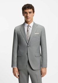 Uomo che indossa un completo grigio chiaro con una camicia bianca, una cravatta a righe argentate e un fazzoletto da taschino a fantasia, in piedi contro uno sfondo bianco semplice.