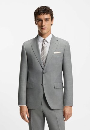Man wearing a light grey suit with a white shirt, striped silver tie, and patterned pocket square, standing against a plain white background.